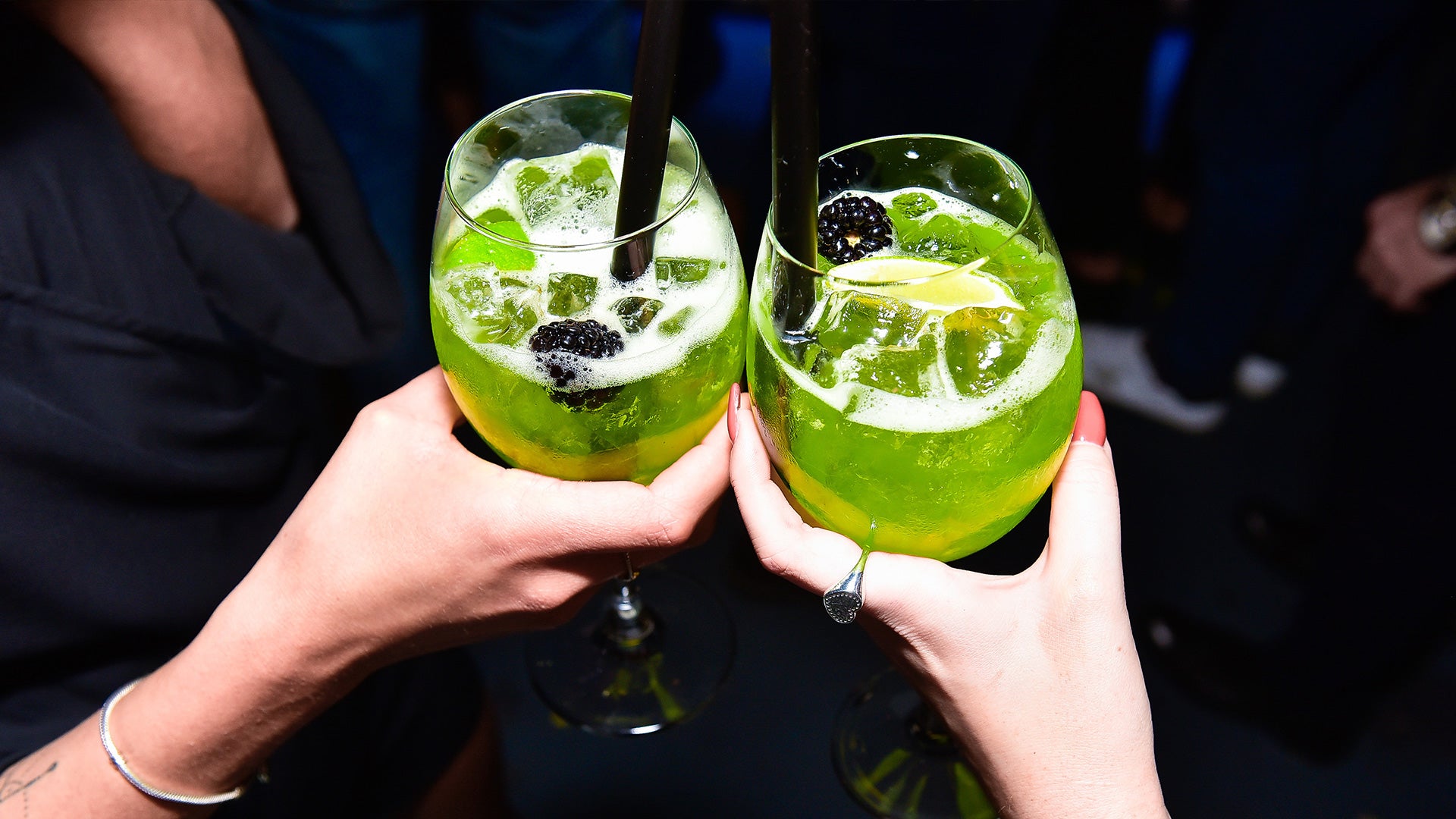 Two hands clinking glasses of green cocktails with ice and blackberries against a dark background