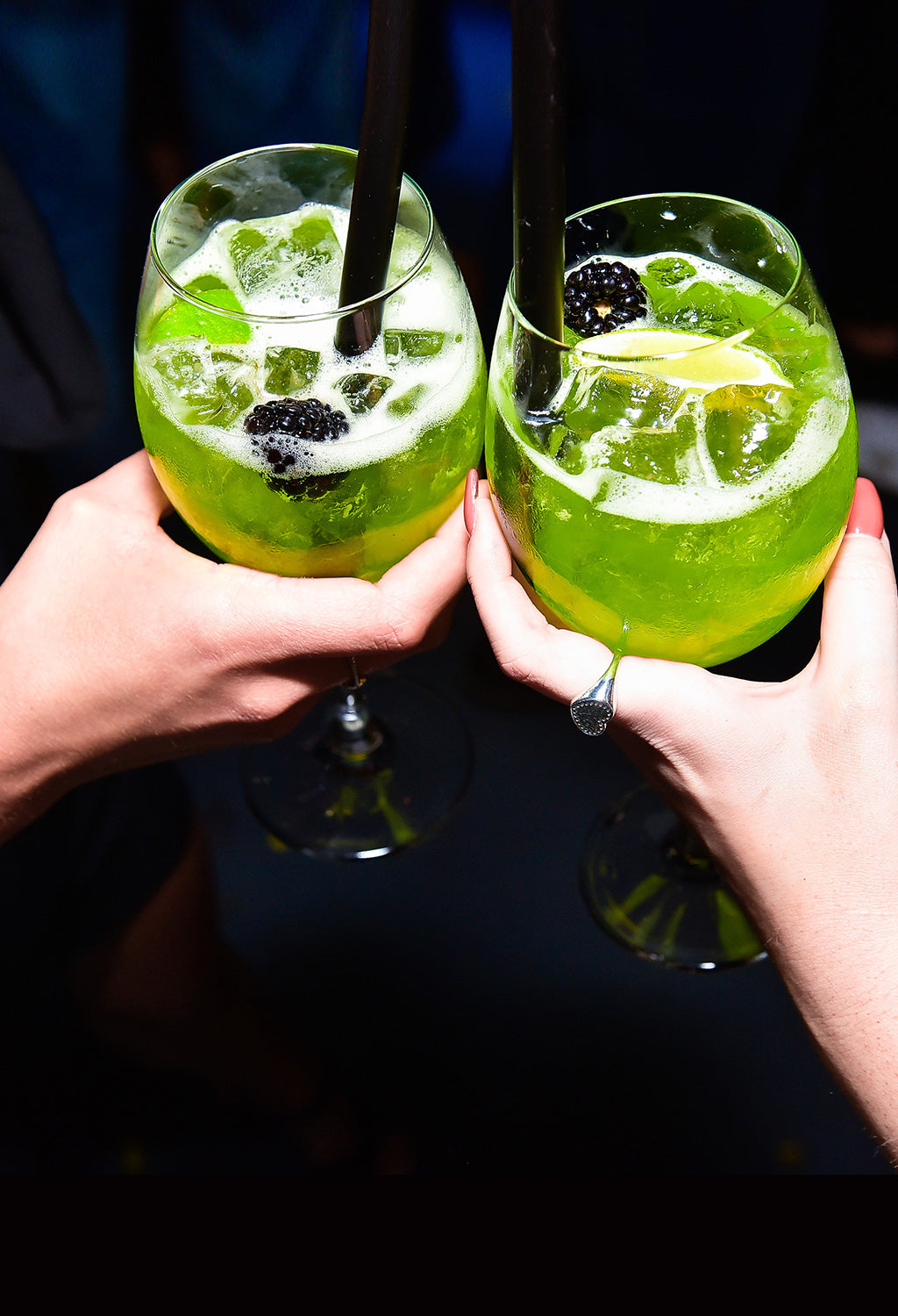 Two hands holding green cocktails with blackberries and lemon slices against a dark background